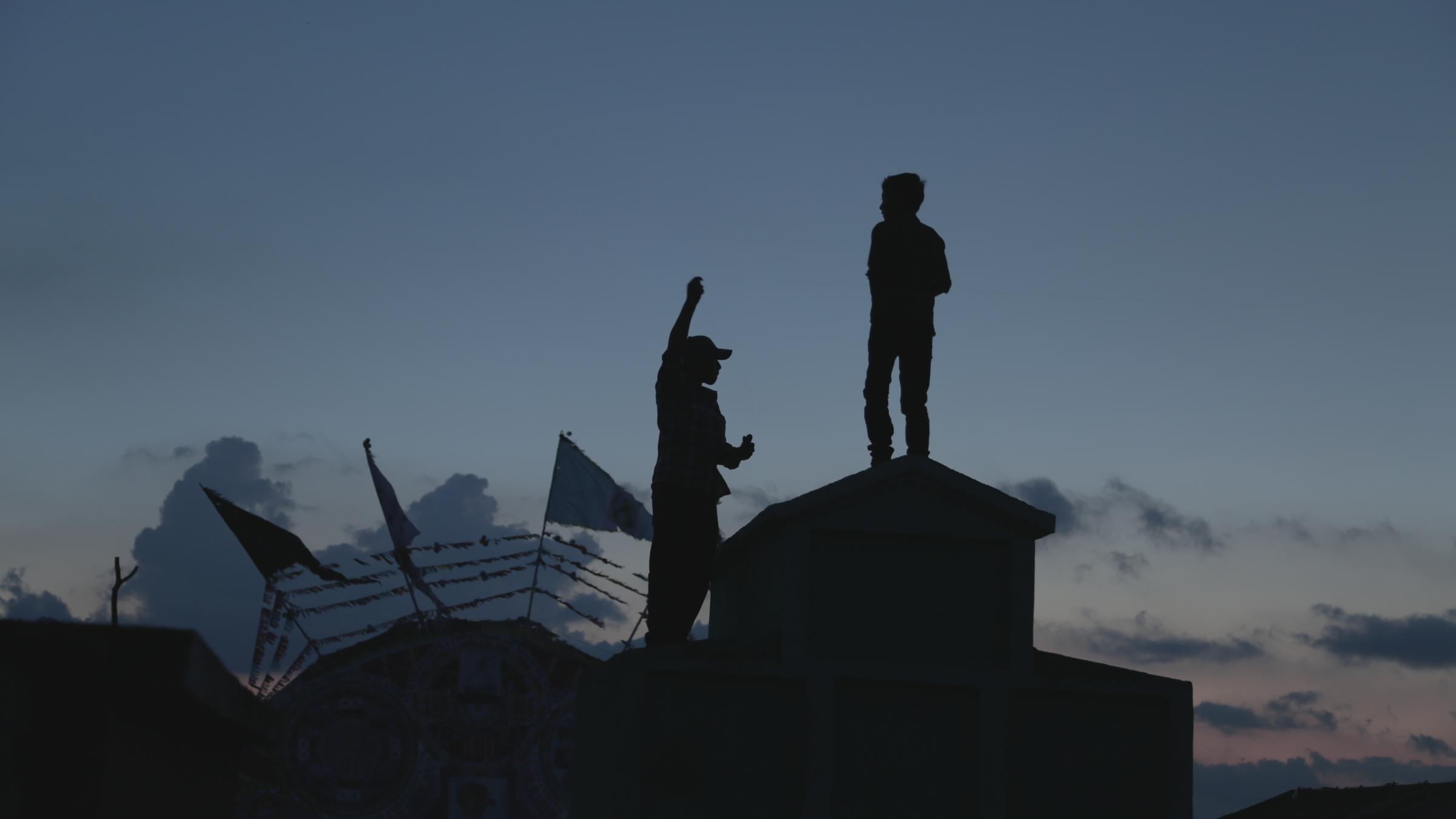 Two figures stand on a rooftop at dusk, flying kites above a cemetery