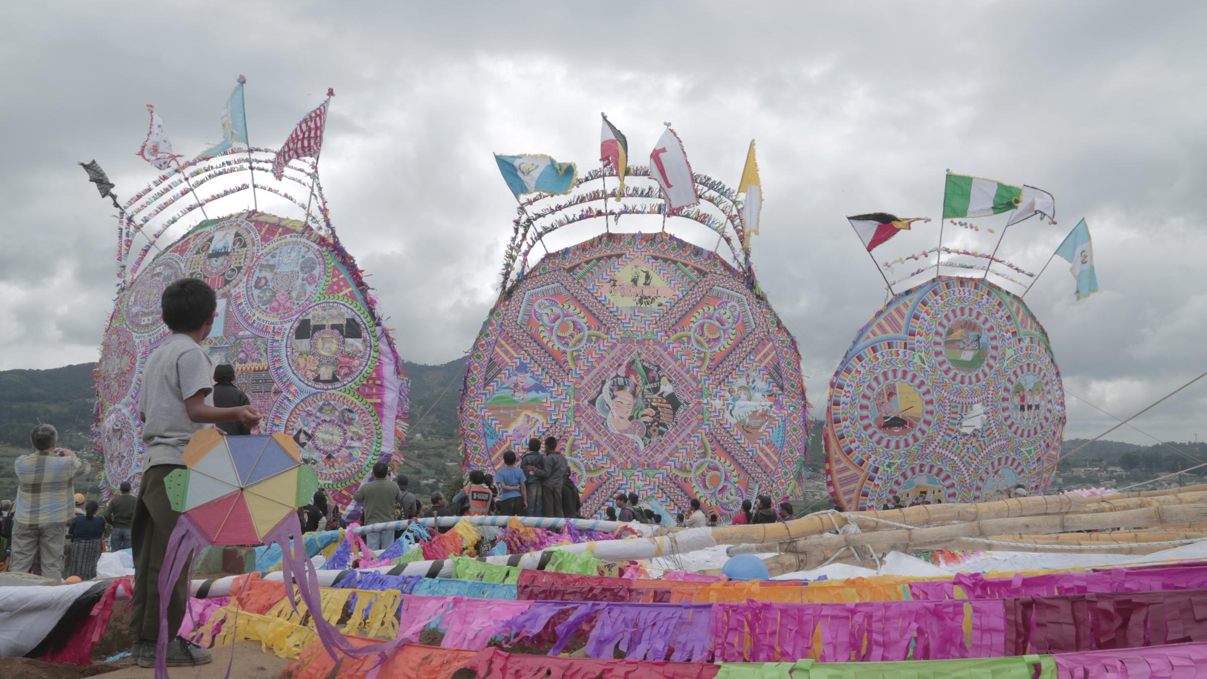 Giant ceremonial kites rise above a hillside cemetery under a clouded sky