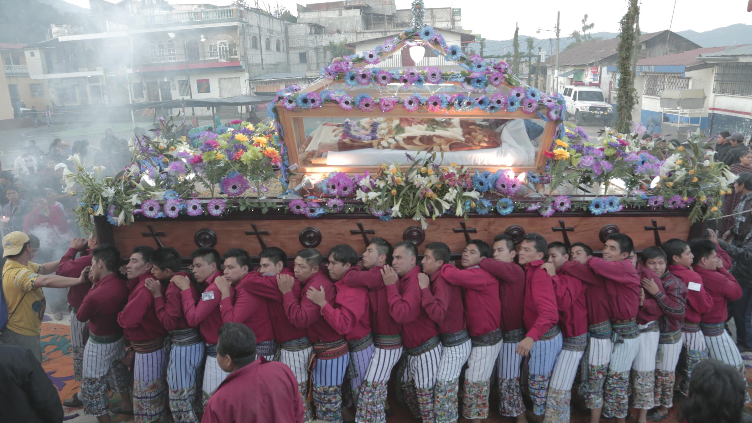 Dozens of men in matching dress carry a flower-covered processional float through incense smoke