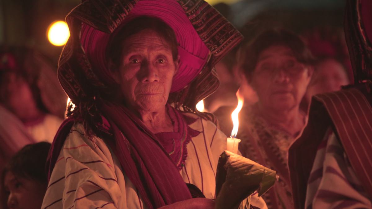 An elder woman in a woven headdress holds a candle during a night procession
