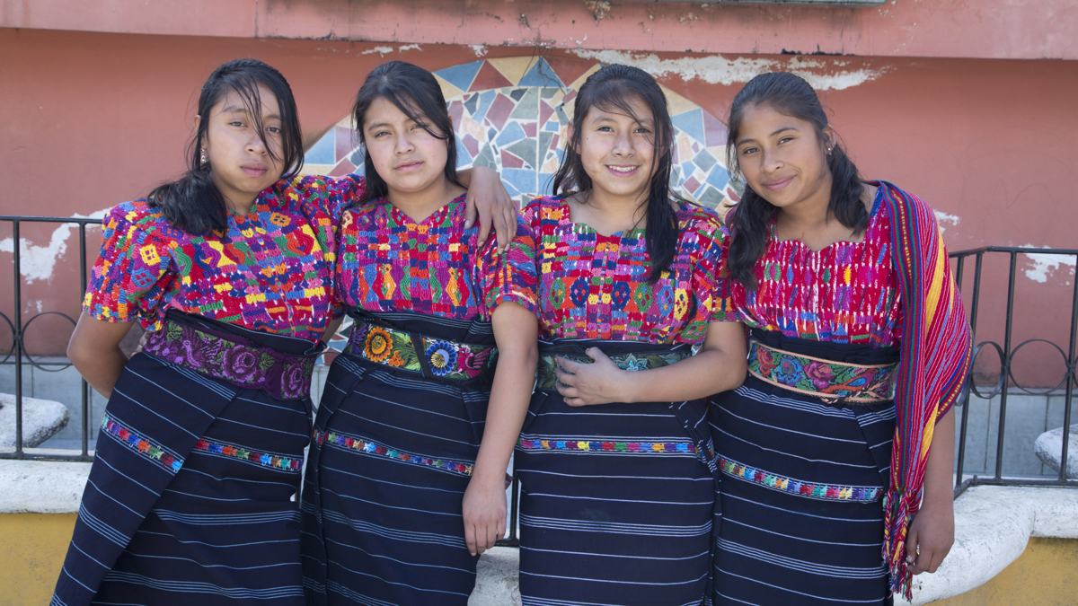 Four young women in embroidered huipiles stand together against a painted wall