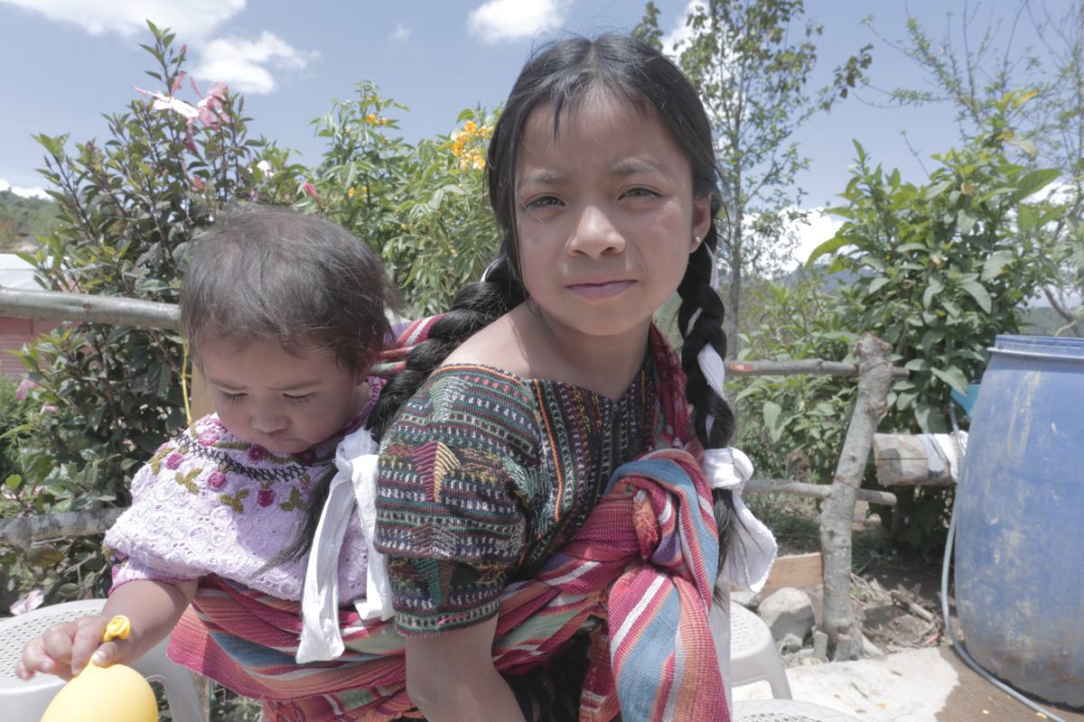 A girl carries her baby sister on her back in a woven shawl