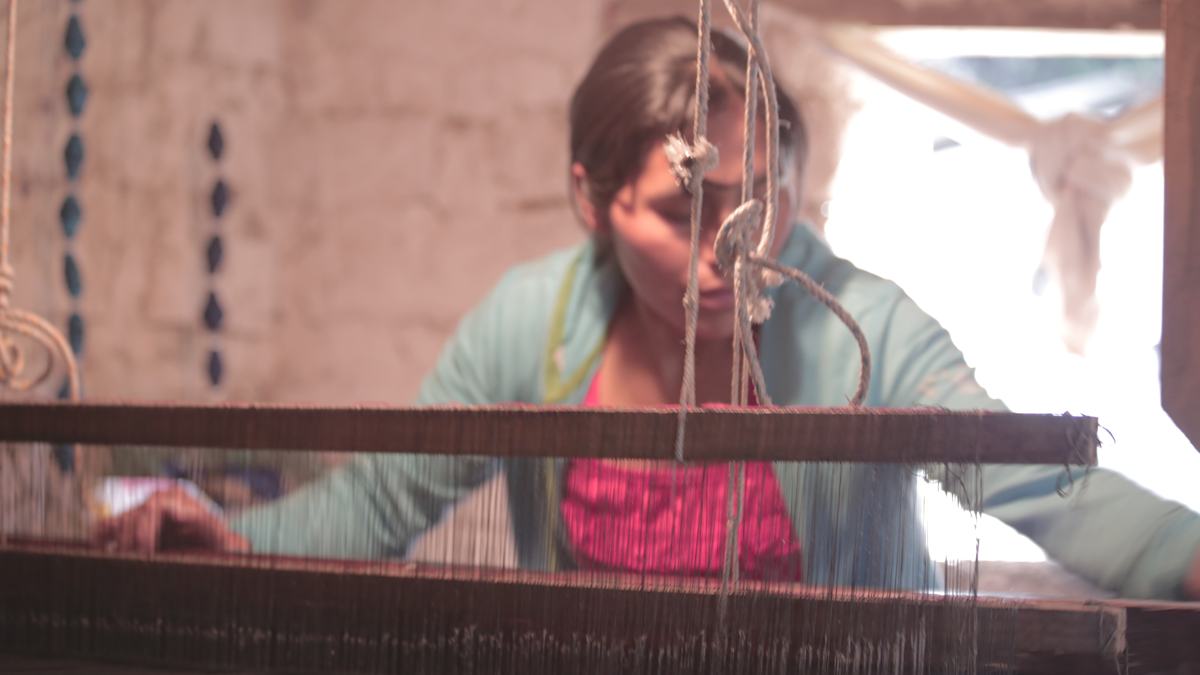 A woman works at a wooden loom, seen through its threads
