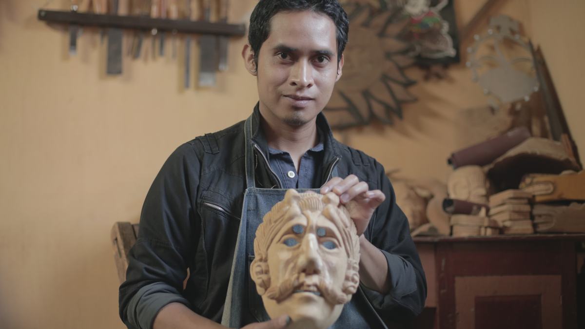 A mask carver holds a carved wooden mask in his workshop