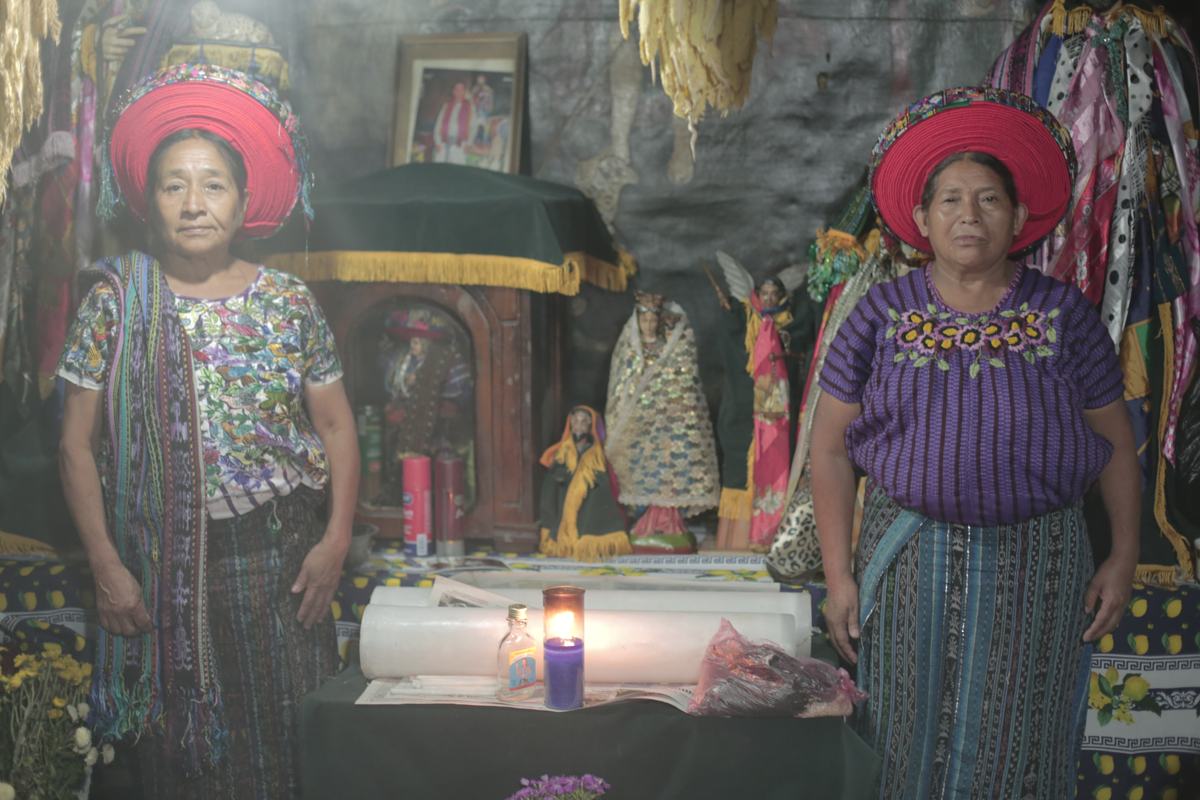 Two women in red headwraps stand before an altar of saints and candles
