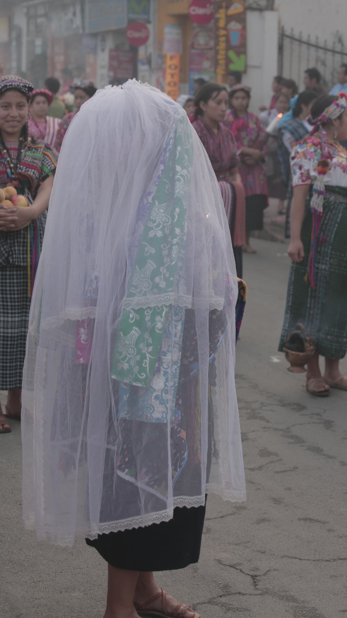 A woman draped in a white veil stands in a procession in the street